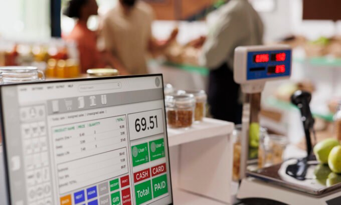 While multiethnic clients are assisted by salesman in the background, apples are placed on digital measuring scale. Photo focus on cashier's desk with weighing equipment and desktop computer.